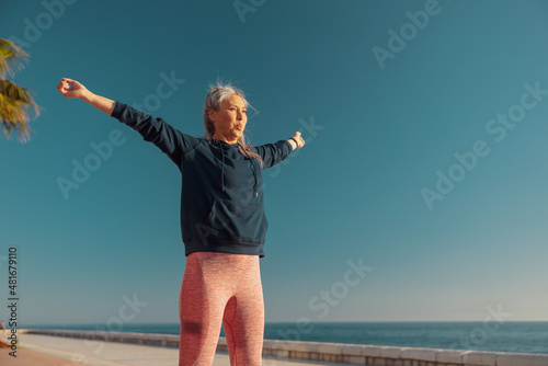 Active woman in a sportswear raising arms to the sides  training and looking ahead by the waterfront