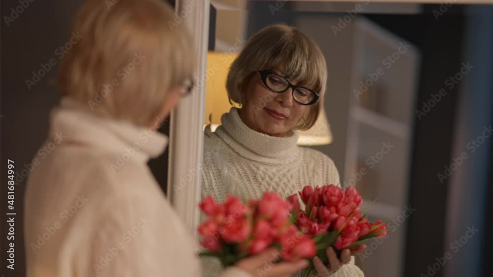 Reflection in mirror of senior woman with bouquet of flowers standing indoors. Beautiful female Caucasian retiree admiring red tulips in living room at home. Birthday or Women's Day celebration Reflection in mirror of senior woman with bouquet of flowers standing indoors. Beautiful female Caucasian retiree admiring red tulips in living room at home. Birthday or Women's Day celebration
