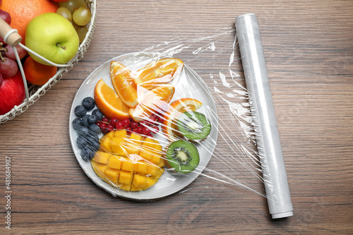 Fototapeta Naklejka Na Ścianę i Meble -  Plate of fresh fruits and berries with plastic food wrap on wooden table, flat lay