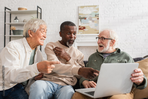 Excited african american man pointing at laptop near multiethnic friends at home.