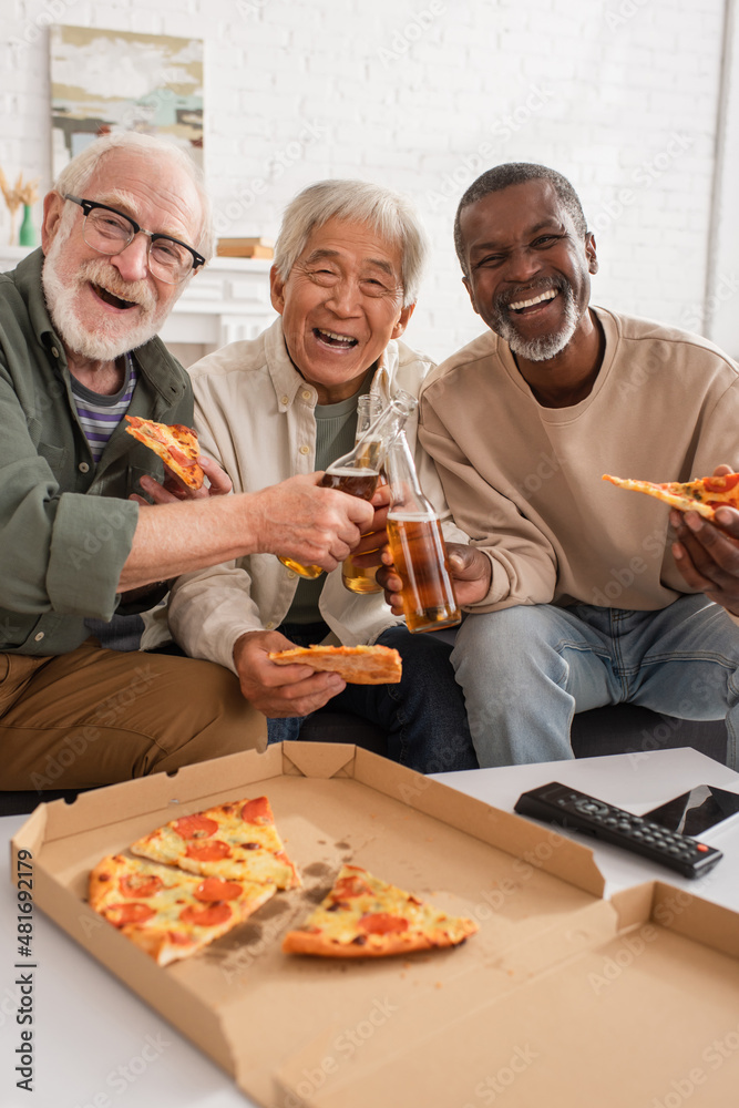 Happy interracial senior men holding pizza and beer near remote controller at home.