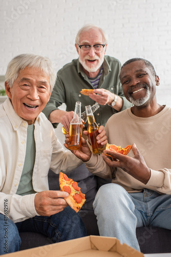 Interracial senior men looking at camera while holding pizza and beer at home.