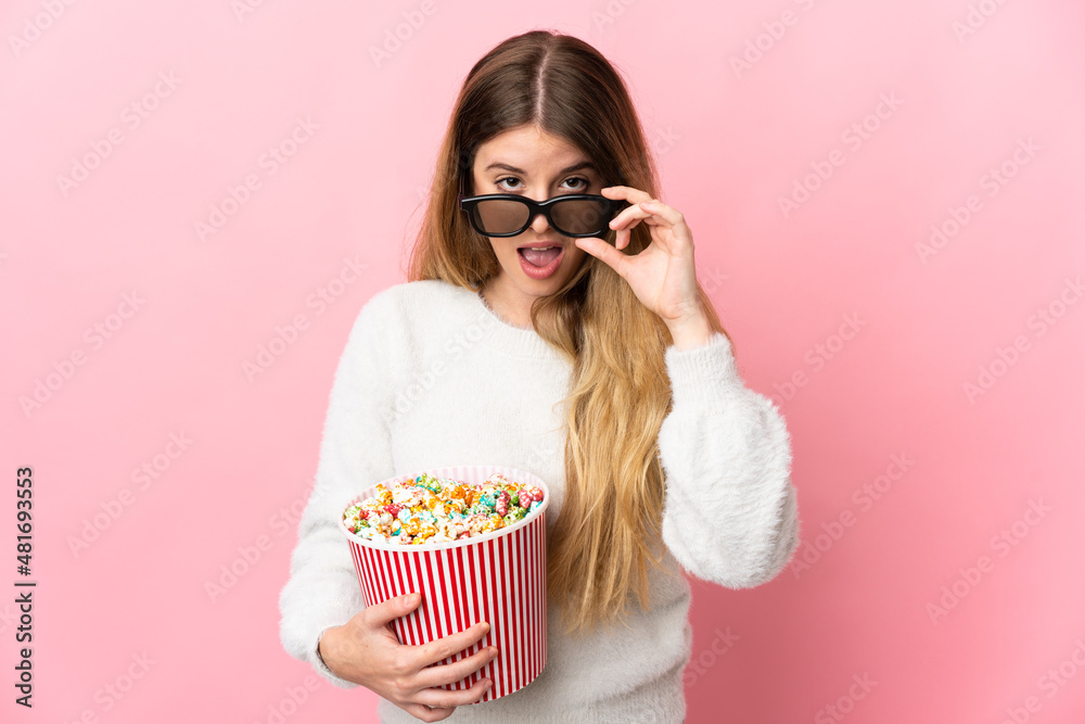 Young blonde woman isolated on pink background surprised with 3d glasses and holding a big bucket of popcorns