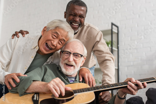 Happy senior interracial friends playing acoustic guitar and looking at camera at home.