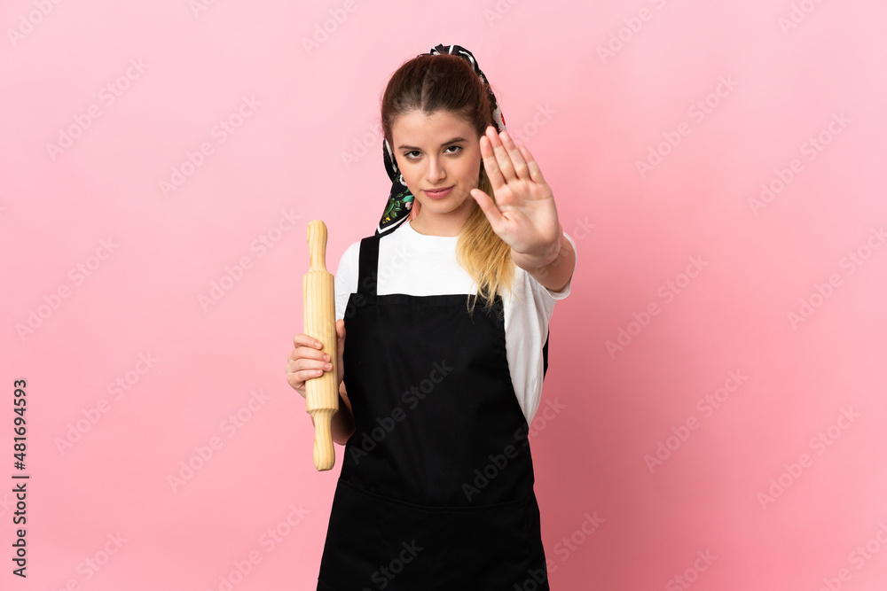 Young blonde woman holding a rolling pin isolated on pink background making stop gesture