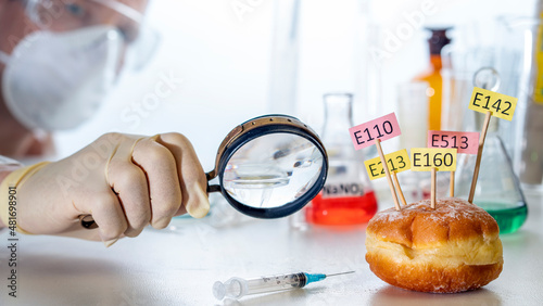 In the laboratory assistant's hand is a magnifying glass, on the laboratory table is a donut decorated with tablets with the names of E. Food Laboratory additives. Healthy food concept.