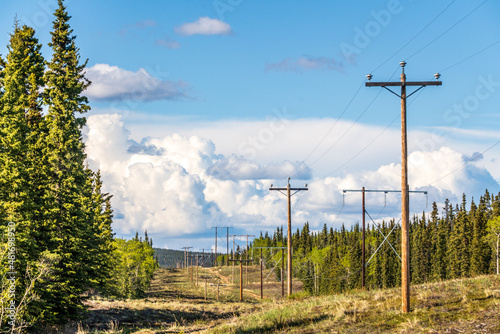 Electricity, power lines seen in rural Canada during summertime with blue sky, clouds and mountains, boreal forest landscape. 