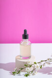 Glass dropper bottle with serum or oil on podium with blooming twigs on pink background. Vertical stock photo with trendy hard shadows