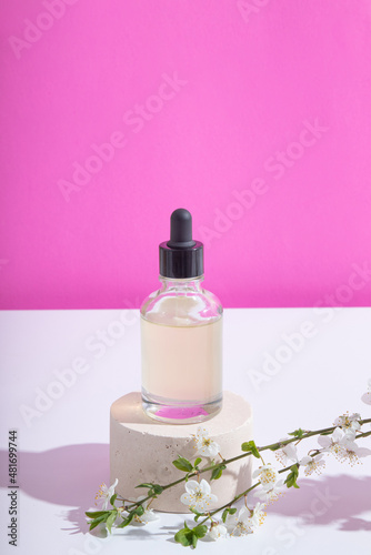 Glass dropper bottle with serum or oil on podium with blooming twigs on pink background. Vertical stock photo with trendy hard shadows