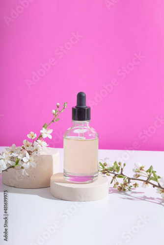 Glass dropper bottle with cosmetic oil on podium with blooming twigs on pink background. Vertical stock photo