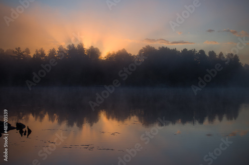early morning sunrise with orange reflecting onto calm waters of cottage lake trees reflecting on tranquil waters suns rays streaming from behind forest religious effect or feel horizontal format 