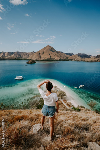 A young female traveler is exploring tropical island in Komodo, Indonesia. She is wearing a casual outfit and overlooks lagoon bag, boat and hills in the distance © Michal