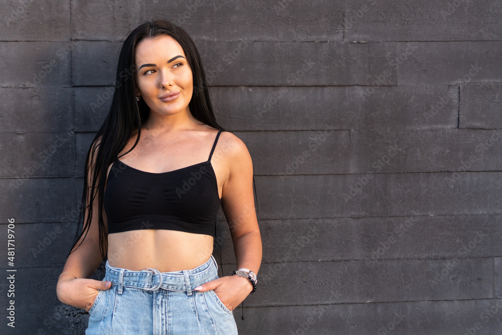 Young woman looking away while posing with her hands in her pockets outdoors against a gray wall. Young woman looking away while posing with her hands in her pockets outdoors against a gray wall.