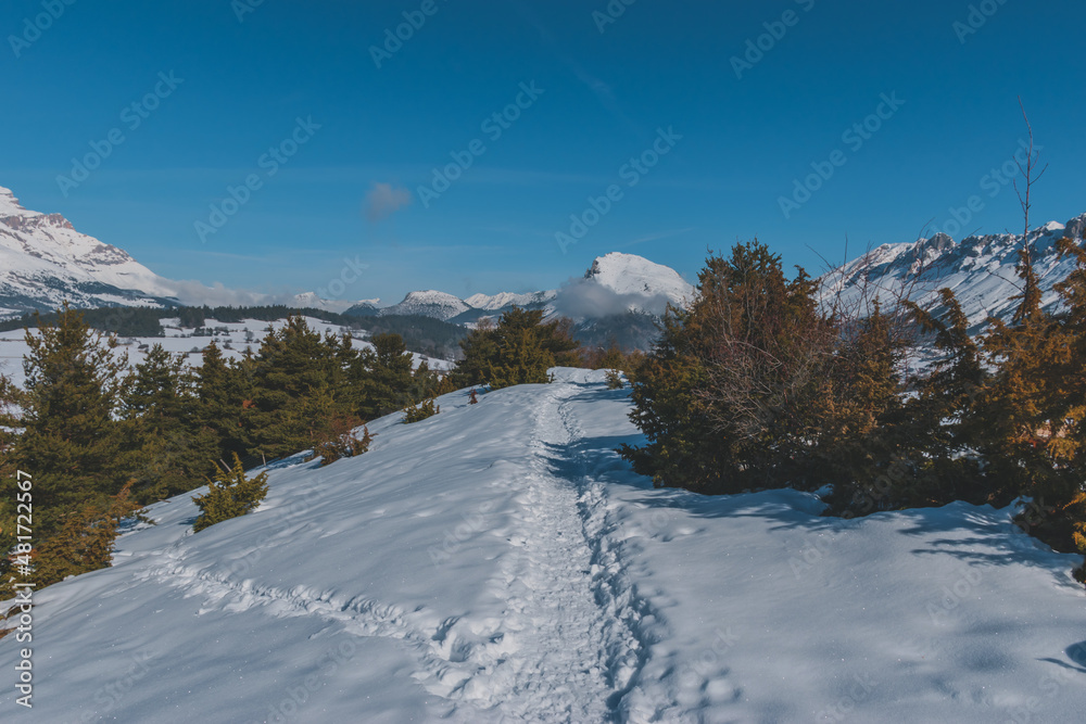 A picturesque landscape view of the snowcapped French Alps mountains with a hiking path in the snow on a cold winter day (La Joue du Loup, Devoluy)