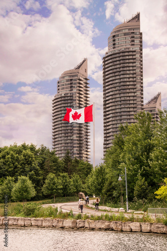 Red white Canadian flag waving on mast above park trees and walking trail in front of modern high-rise buildings of Humber Bay Shores, historic and scenic part of Etobicoke district of Toronto City photo