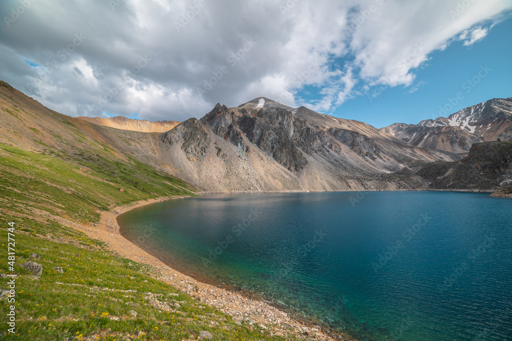 Deep mountain lake of phantom blue color among high mountains with pointed peak in changeable weather. Wonderful dramatic view to deep blue mountain lake among sunlit sharp rocks under cloudy sky.