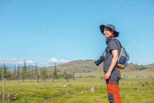 One traveling man with hat and backpack walks in sunlight on vivid bokeh background of coniferous trees and mountain under blue sky. Colorful portrait of tourist with camera in bright sun in mountains