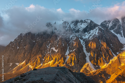 Scenic landscape with snowy mountain top in low clouds in golden sunrise colors. Colorful view to snow mountains and rocks in gold morning sunlight in low clouds. Awesome scenery with golden rocks.