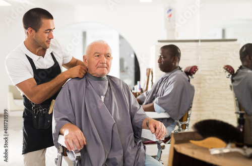 Portrait of young friendly barber putting on hairdresser cloak to elderly male client sitting in haircut chair in salon