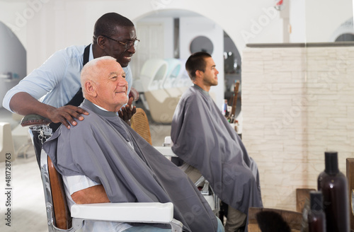 Positive gray-haired male client sitting in hairdressing chair, discussing haircut with African hairdresser