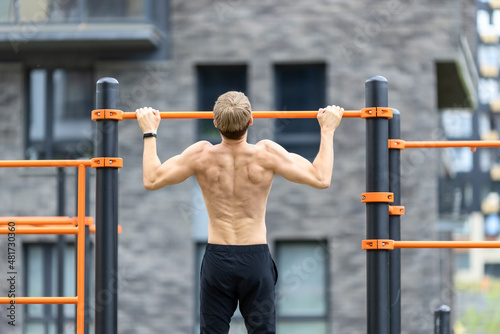Shirtless muscular man with fit torso having outdoor fitness workout on pull up bar in the street open gym