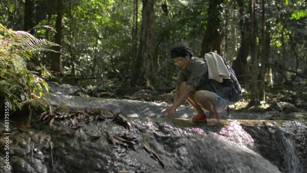 Asian female backpacker washing her hands in the cascade that flowing over the rocks among lush foliage vegetation in rainforest during summer trip. Solo camping. Travel explorer in the nature. Asian female backpacker washing her hands in the cascade that flowing over the rocks among lush foliage vegetation in rainforest during summer trip. Solo camping. Travel explorer in the nature.