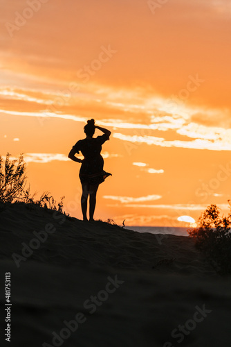 Unidentifiable woman stands on sand dunes by the seaside watching the sunset. Silhouette of woman in dress.