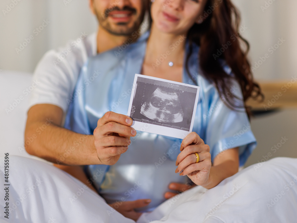 Cheerful husband embracing happy wife on bed at night and showing pregnancy ultrasound image film of unborn baby Cheerful husband embracing happy wife on bed at night and showing pregnancy ultrasound image film of unborn baby