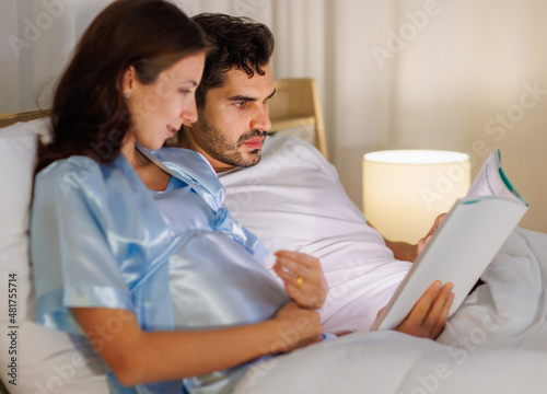 Happy pregnant wife holding belly and relax on bed beside beloved husband and enjoy reading entertainment media on a book together with smile and laugh. Parents expecting joyful life for child birth.