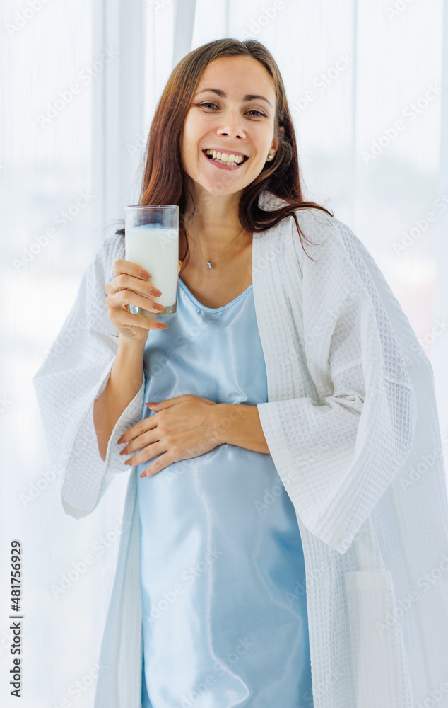 Pregnant woman on night gown happy to drink glass of milk beverage while relaxing near window curtains Pregnant woman on night gown happy to drink glass of milk beverage while relaxing near window curtains