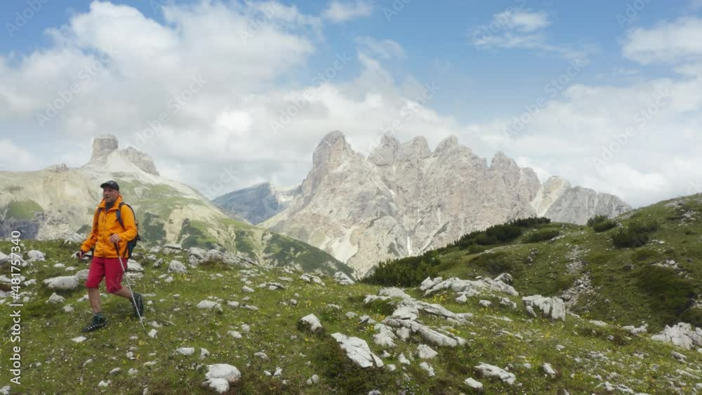 Man trekker walking with backpack and trekking poles by green mountain hill and enjoying picturesque Dolomite Alps range peaks view near Tre Cime in Italy active people and European mountain concept Man trekker walking with backpack and trekking poles by green mountain hill and enjoying picturesque Dolomite Alps range peaks view near Tre Cime in Italy active people and European mountain concept