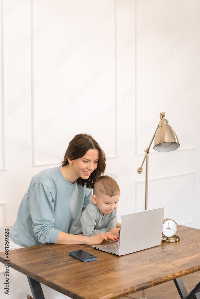 Young mom and son surf on the Internet. A little boy with light blond hair at a table plays on a laptop. Young mom and son surf on the Internet. A little boy with light blond hair at a table plays on a laptop.