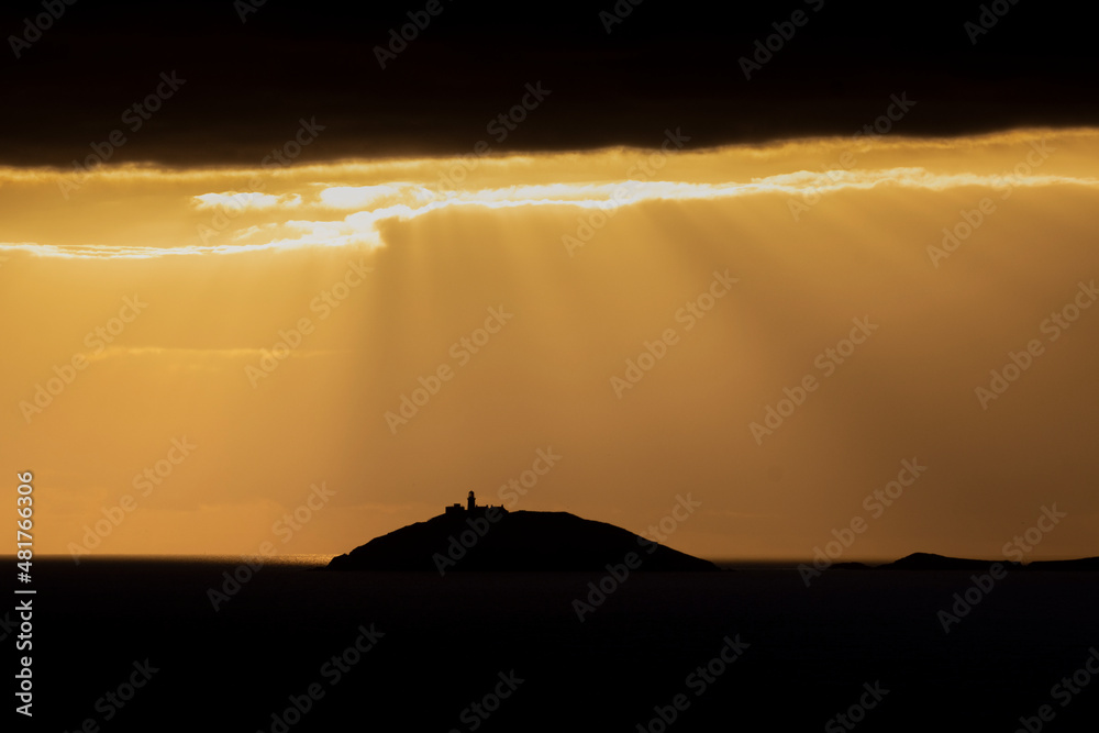 Rays of sun shine through clouds during sunset on a calm winter day with Ballycotton Lighthouse in county Cork, Ireland, in the background