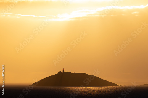 Rays of sun shine through clouds during sunset on a calm winter day with Ballycotton Lighthouse in county Cork  Ireland  in the background