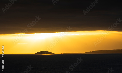 Rays of sun shine through clouds during sunset on a calm winter day with Ballycotton Lighthouse in county Cork  Ireland  in the background