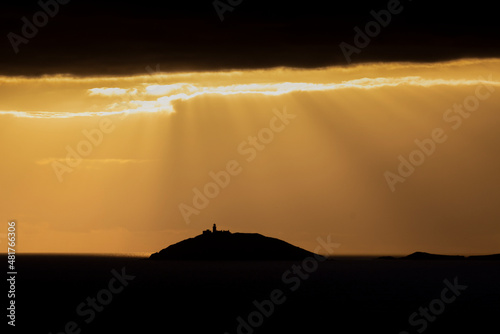 Rays of sun shine through clouds during sunset on a calm winter day with Ballycotton Lighthouse in county Cork  Ireland  in the background