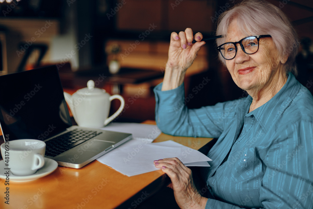 Portrait of an elderly woman with glasses sits at a table in front of a laptop Freelancer works unaltered Portrait of an elderly woman with glasses sits at a table in front of a laptop Freelancer works unaltered