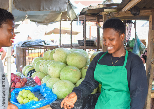 A happy African woman or female trader in green apron trying to sell her goods to a customer at a local market