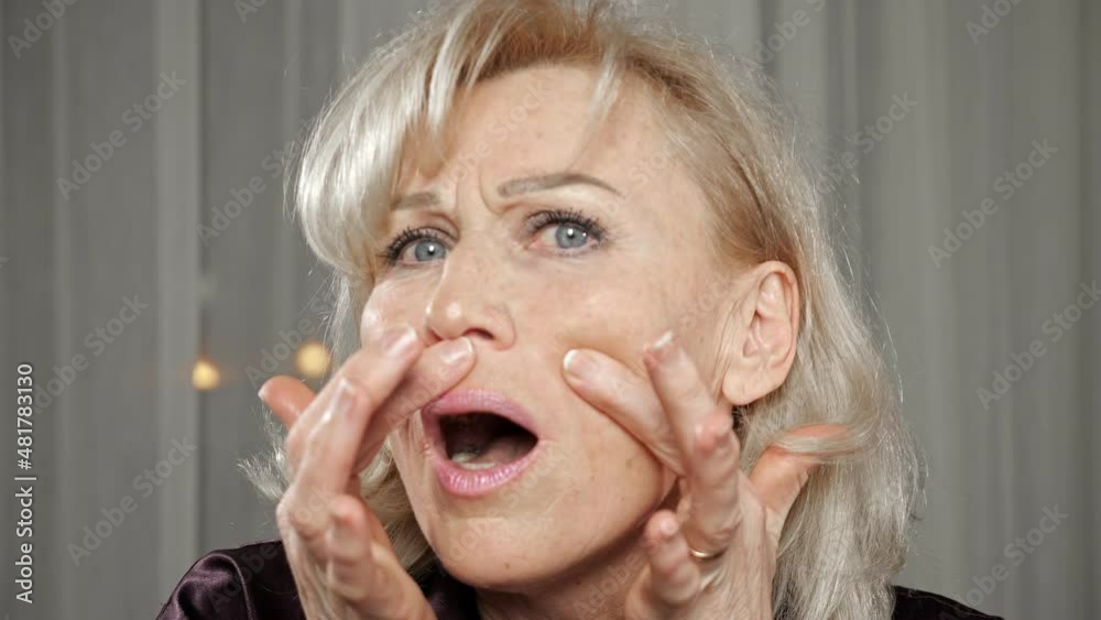 Elderly blonde woman with light natural makeup wearing silk pajamas checks wrinkles on face frowning and looking attentively in mirror closeup.