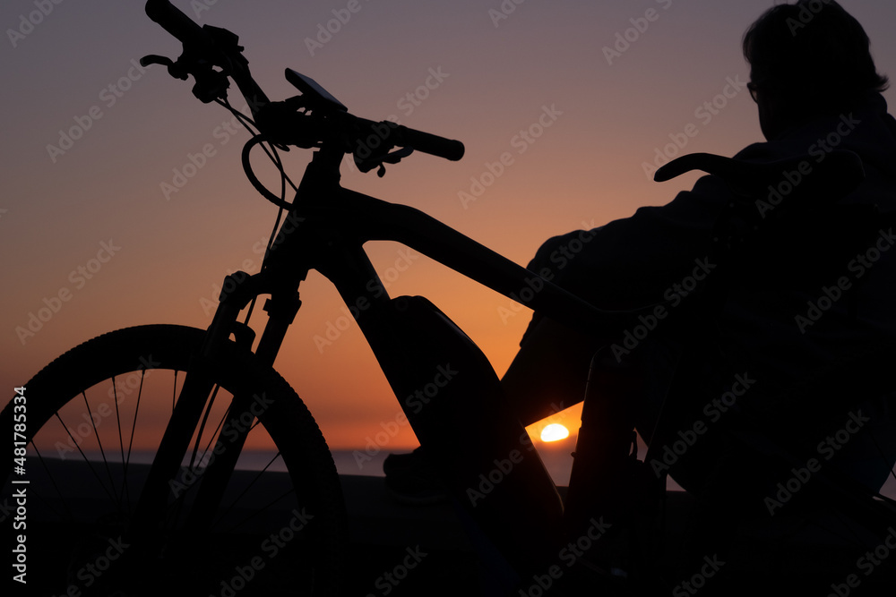 Obraz premium Silhouette of adult woman looking at sunset sitting after cycling along the sea. People enjoying beauty in nature. Golden orange sunset over water