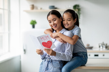 Happy asian daughter greeting mom with Mother's day and hugging her, sitting on dinner table at kitchen interior