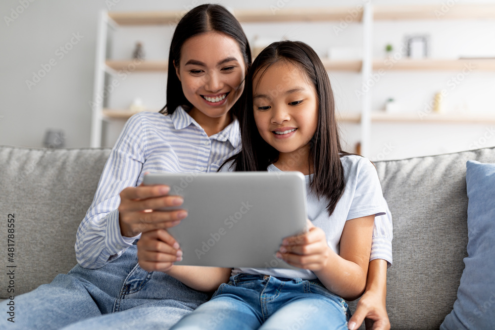 Happy asian mother and child girl using digital tablet together while sitting on couch at home, free space