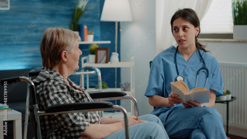 Obraz premium Medical assistant reading book for elder disabled woman in nursing home. Senior patient sitting in wheelchair receiving assistance and support from nurse as entertainment and fun activity.