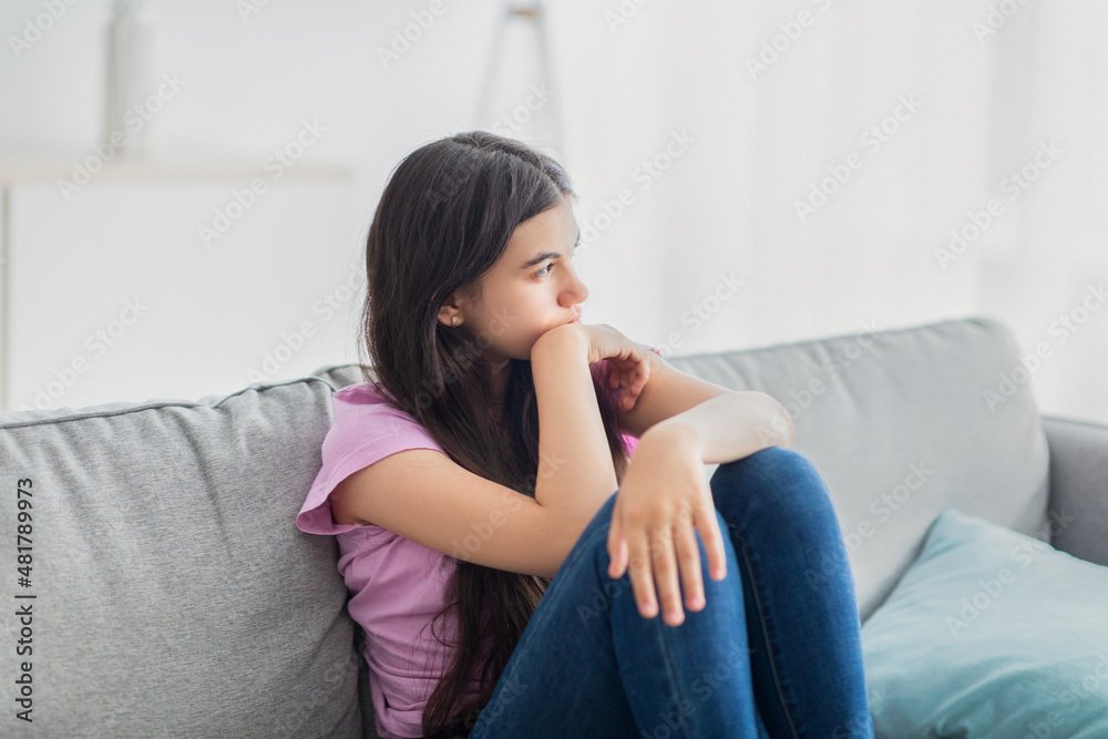 Stressed Indian teenage girl sitting on sofa at home, feeling upset and lonely, suffering from lack of communication Stressed Indian teenage girl sitting on sofa at home, feeling upset and lonely, suffering from lack of communication