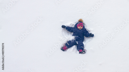 A baby girl lies in the snow in winter and waves her hands portraying an angel
