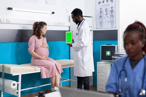 Pregnant patient looking at vertical green screen on tablet in office. Medical physician holding chroma key on device display for mockup template and isolated background at checkup visit.