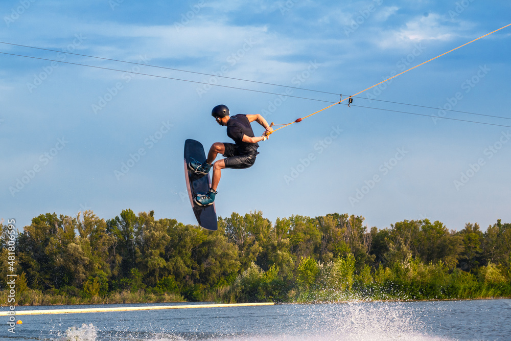 Skilled male wakeboarder jumping with backside rotation over water holding rope in hand