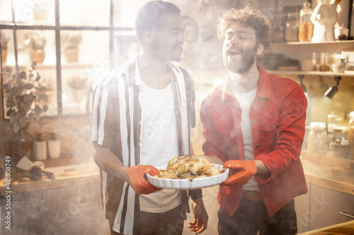 Portrait of two lovely multiracial guys standing together with cooked turkey on kitchen with a steam on background from the oven. Concept of gay relations everyday life together at home
