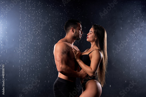 sexy young man gently hugging wet sensual woman in water drops on black background, portrait of caucasian lovely couple with sportive athletic body posing at camera, on love. in rain, water drops