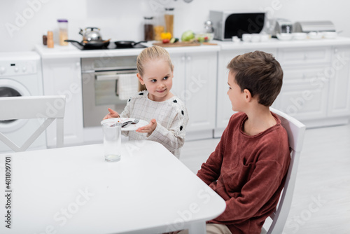 smiling girl holding plate with cutlery near brother sitting at kitchen table.